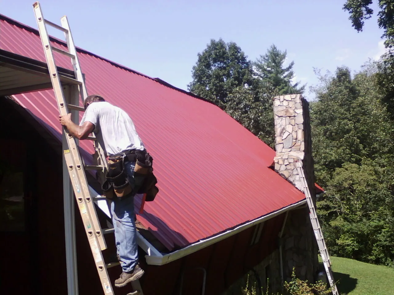 Roofer on a ladder beside a red metal roof and stone chimney — Soffit & Fascia Repair by Roofing Brothers in Grundy Center